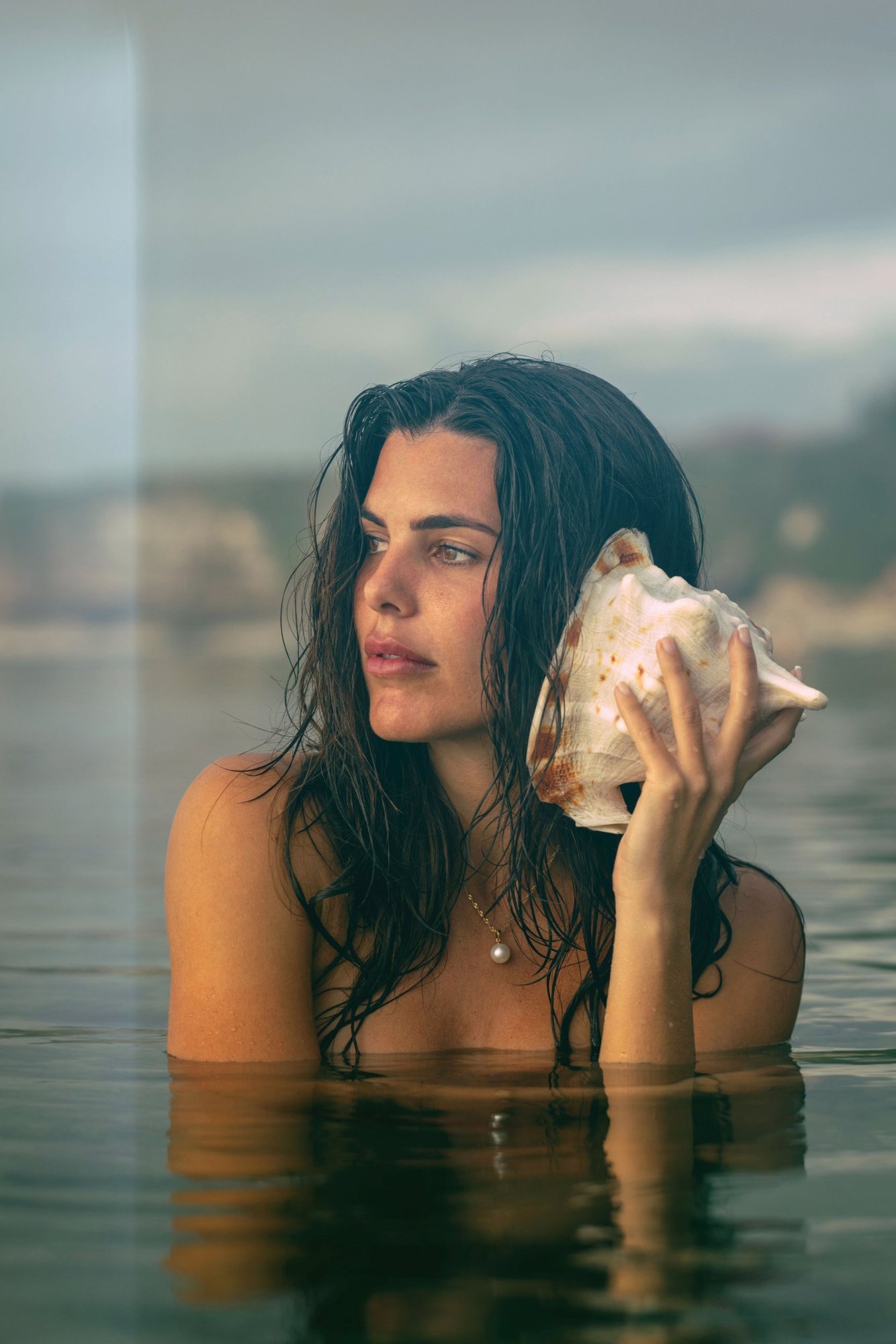 Aquila in calm water at dusk, holding a conch shell to her ear